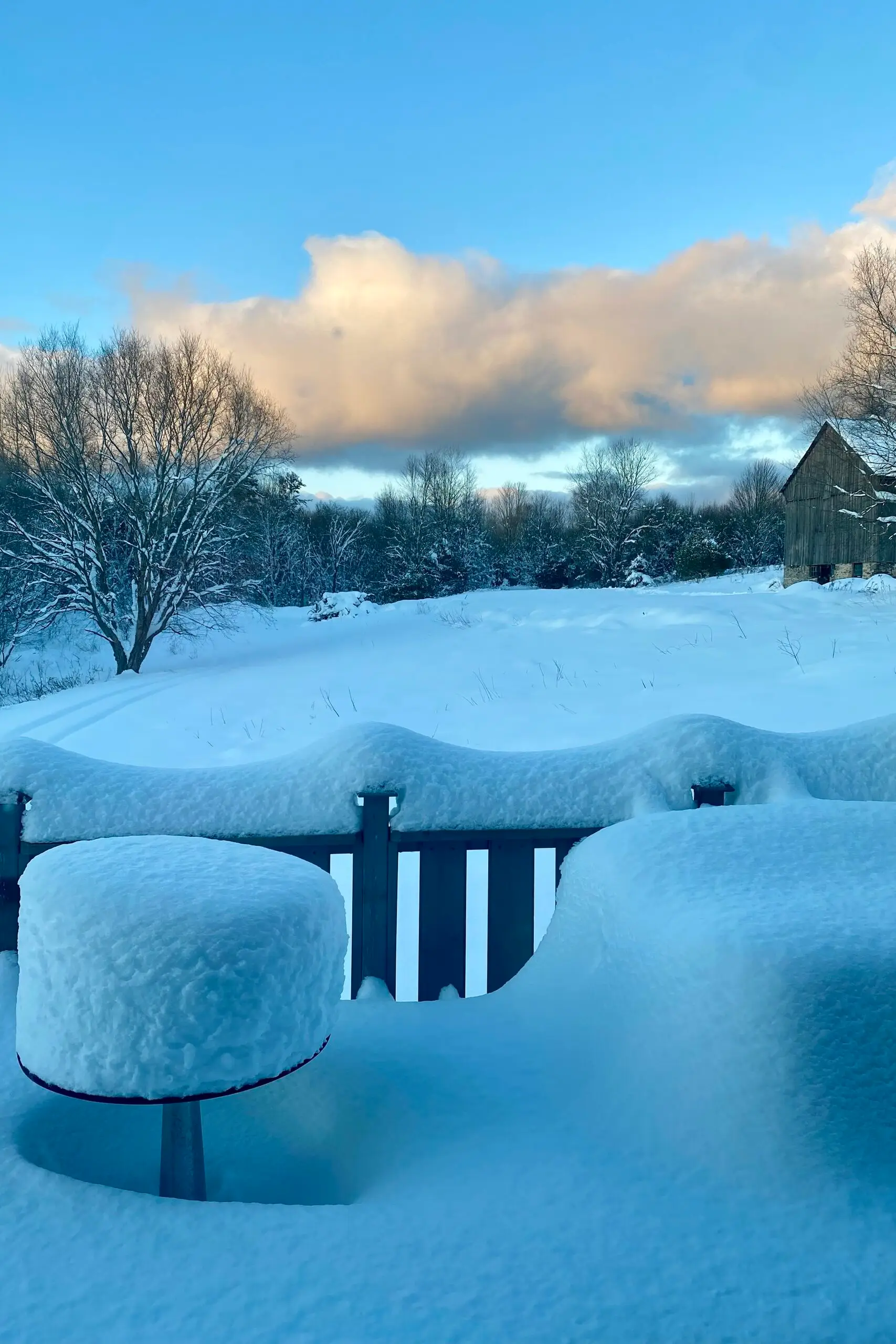 Deep snow piled in smooth drifts along a deck railing, with the barn faint in the winter distance